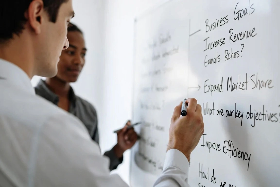 Two colleagues writing business goals and objectives on whiteboard